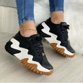 Black and white sneakers with brown soles worn by a person on a light wooden floor.