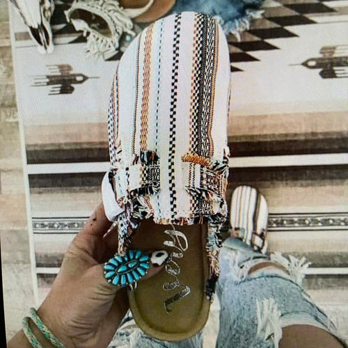 Hand holding a sandal with decorative strap and turquoise charm on a patterned rug background